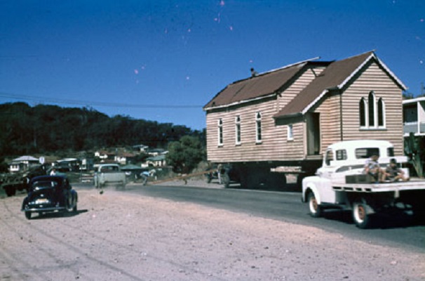 A wood-clad church with archway windows is being transported on an oversize trailer. Cars typical of the 1950s drive on the dirt to avoid it. Two children are riding on the open trailer of a 1960s truck.