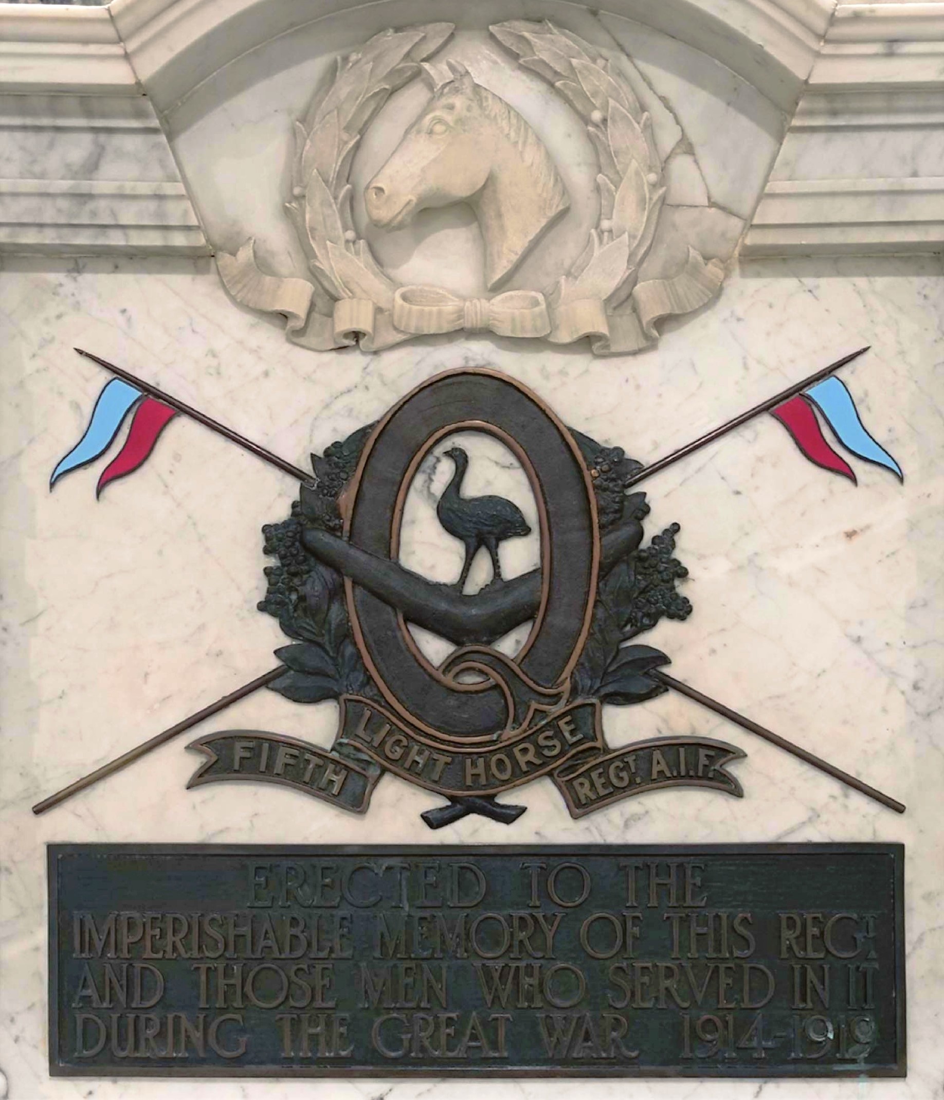 Dark brown plaque of letter Q surrounded by wattle wreath, with emu in centre standing on boomerang, blue and red flags crossing in the background. Words below and white horse head in wreath above.