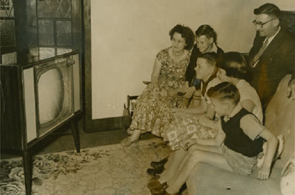 A family dressed in 50s clothes gather around a 50s television, watching from the couch.