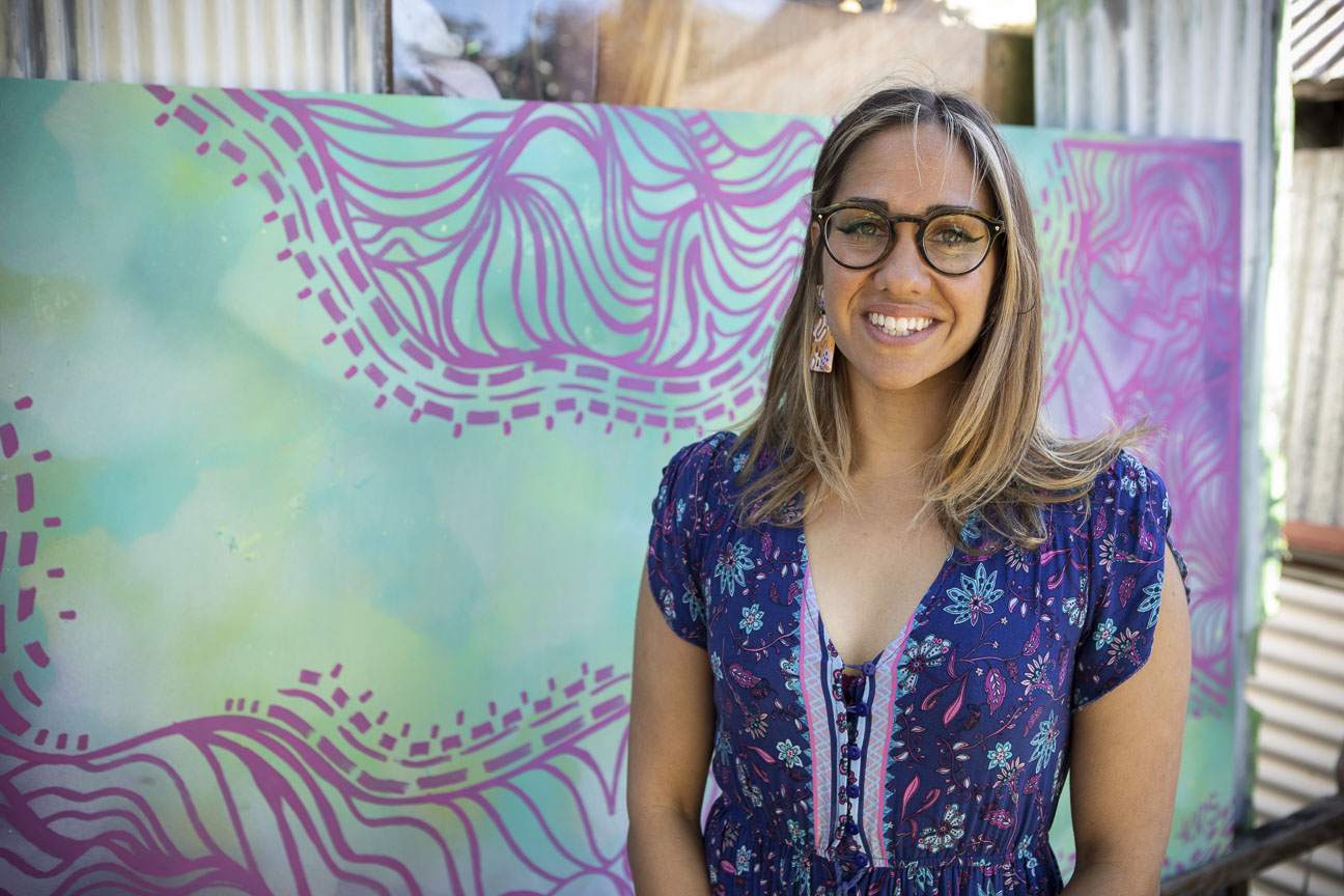 Kirli Saunders stands in a purple top, with glasses, smiling. Behind her is her artwork - it looks green and purple stylised botanical