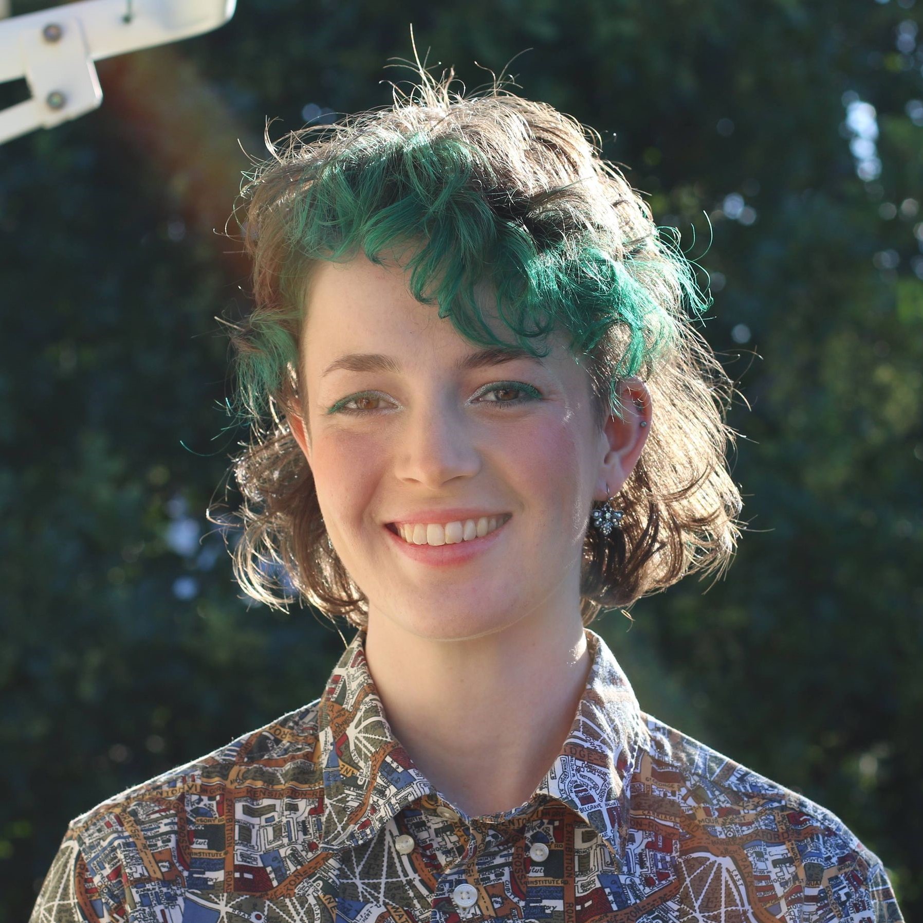 Ellie Kaddatz stands smiling outside in the sun. Her hair is coloured green at the fringe and she wears a patterned shirt. 