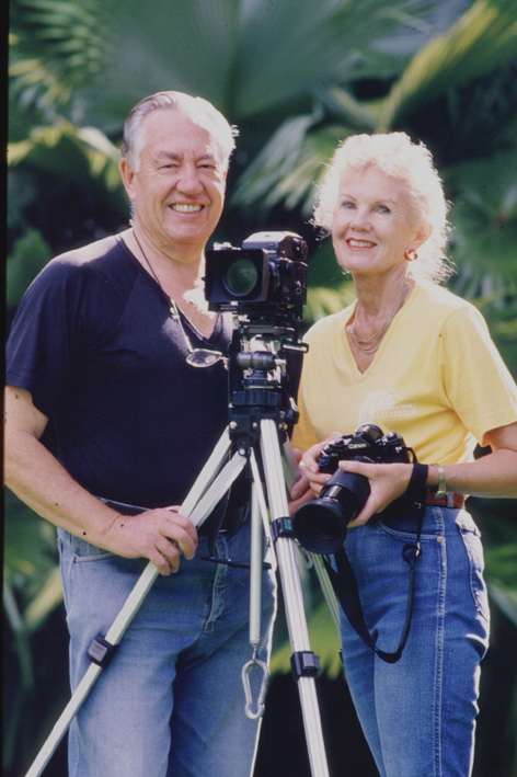 Photographers Ron and Ngaire pose with their Cameras infront of a palm-green background