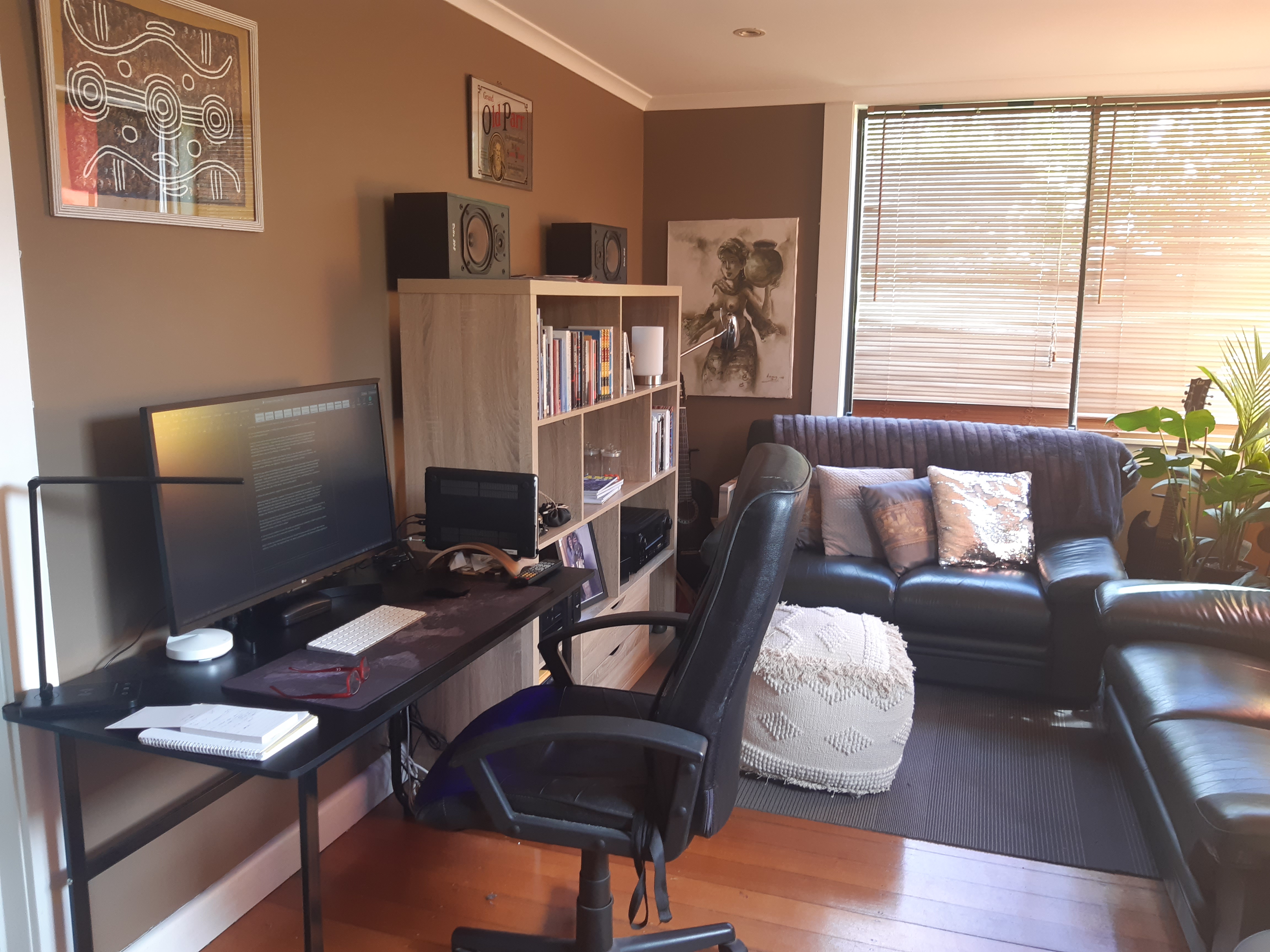 An image of a home office with a black desk and chair, a big computer monitor, and couches, bookshelves and cushions around it.