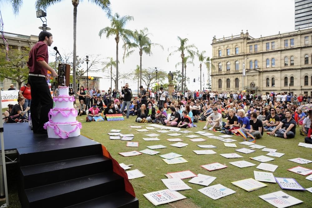 National day of action for marriage equality rally in Brisbane. A person stands on a stage next to a wedding cake made of paper. They are speaking into a microphone to a large crowd sat on the grass carrying rainbow flags and signs.
