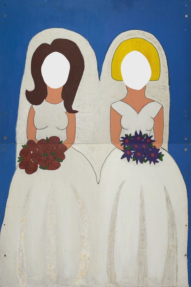 A painted cardboard cutout of two women in bridal gowns holding wedding bouquets. The faces are cut out so people can put their heads through.