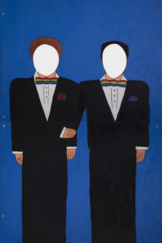 A painted cardboard cutout of two men in groom’s suits with rainbow bow ties. The faces are cut out so people can put their heads through.