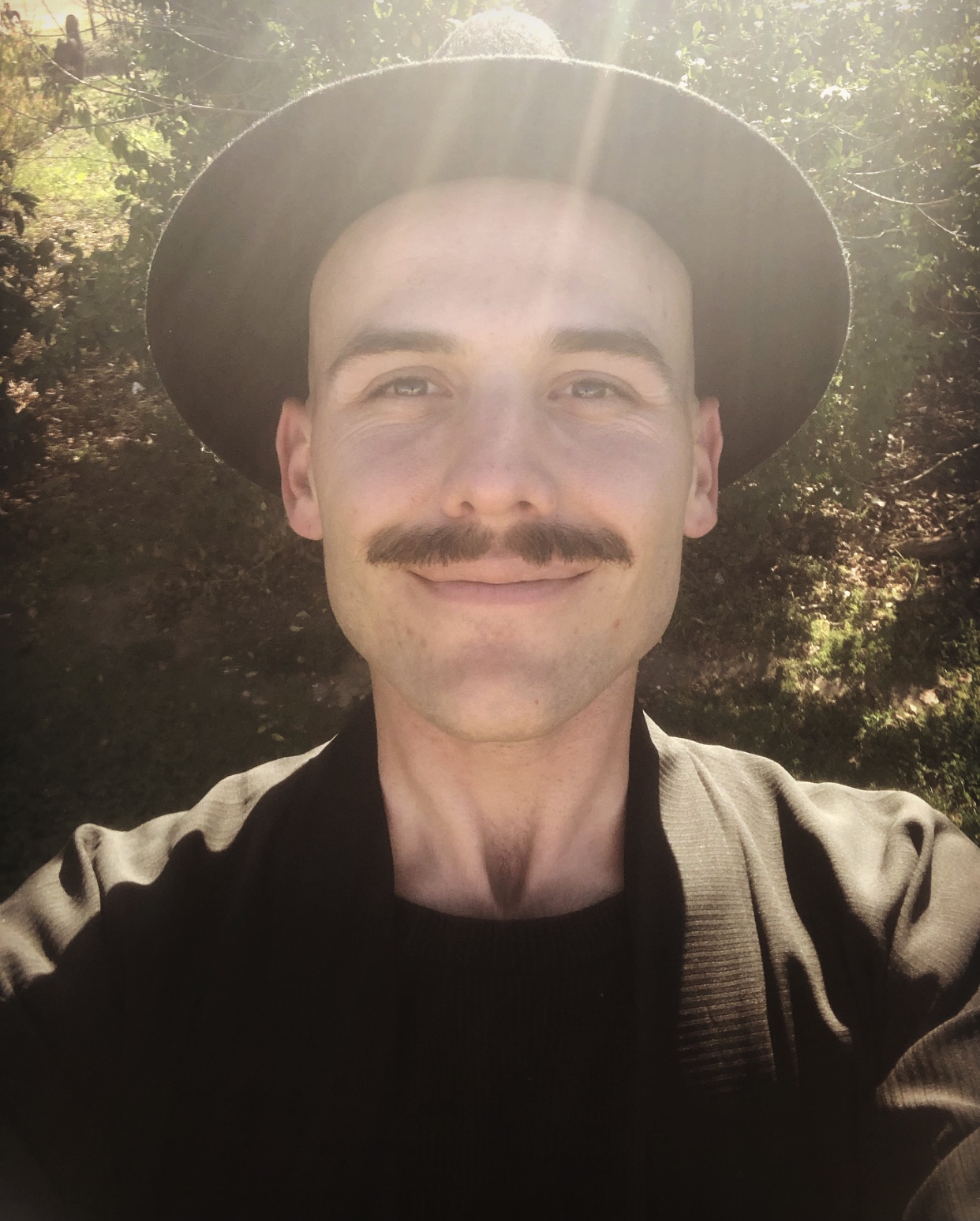 A selfie of Darby Jones, who is wearing a black jacket and hat. He is smiling at the camera with sun and a tree behind him.