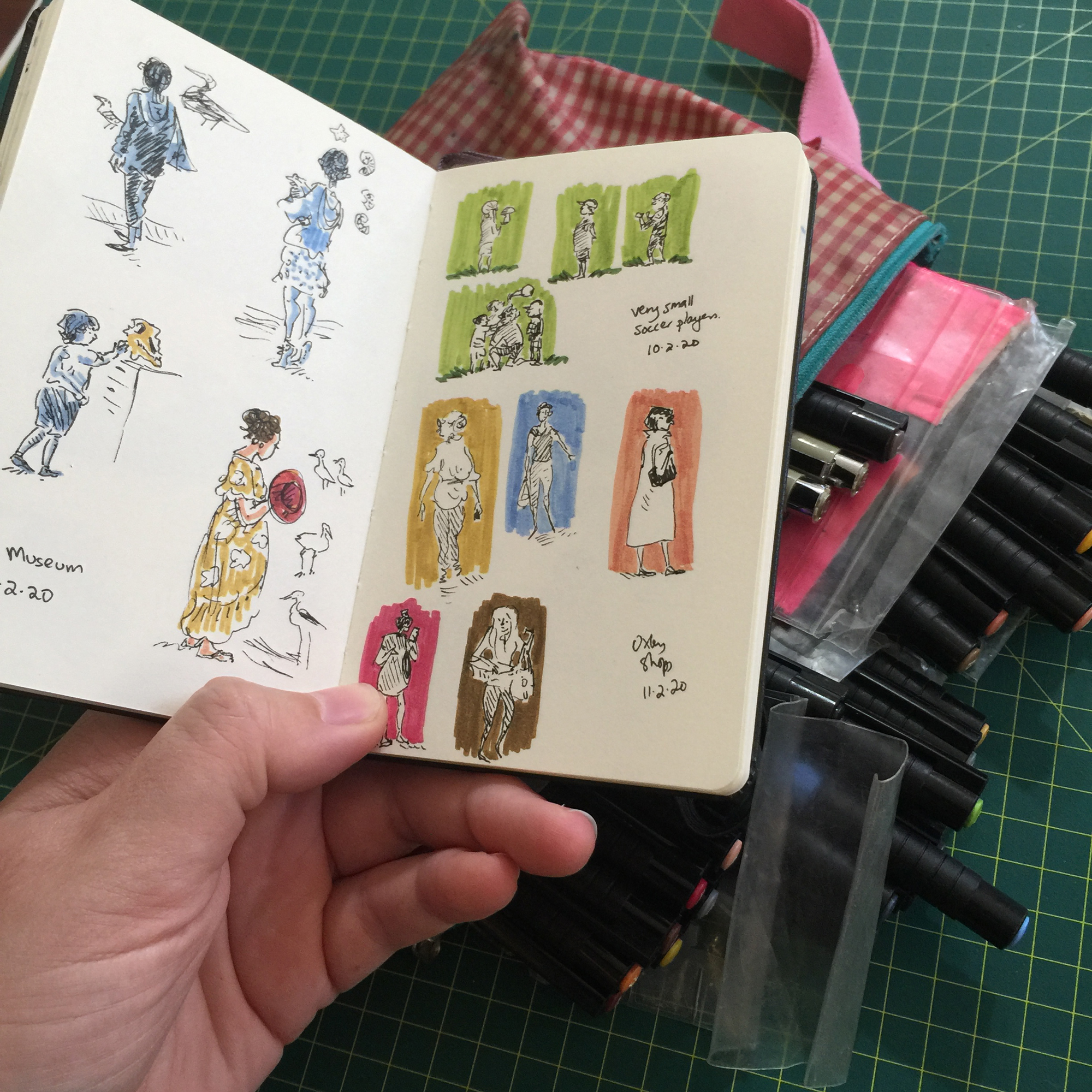 An open sketchbook held in front of a collection of illustrator's pens
