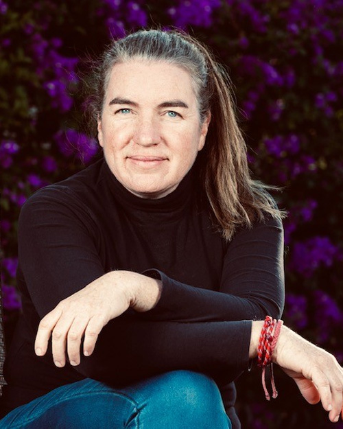 Photo of A E Macleod. They are seated with arms crossed over, wearing a black top, and in front of a purple flowered background