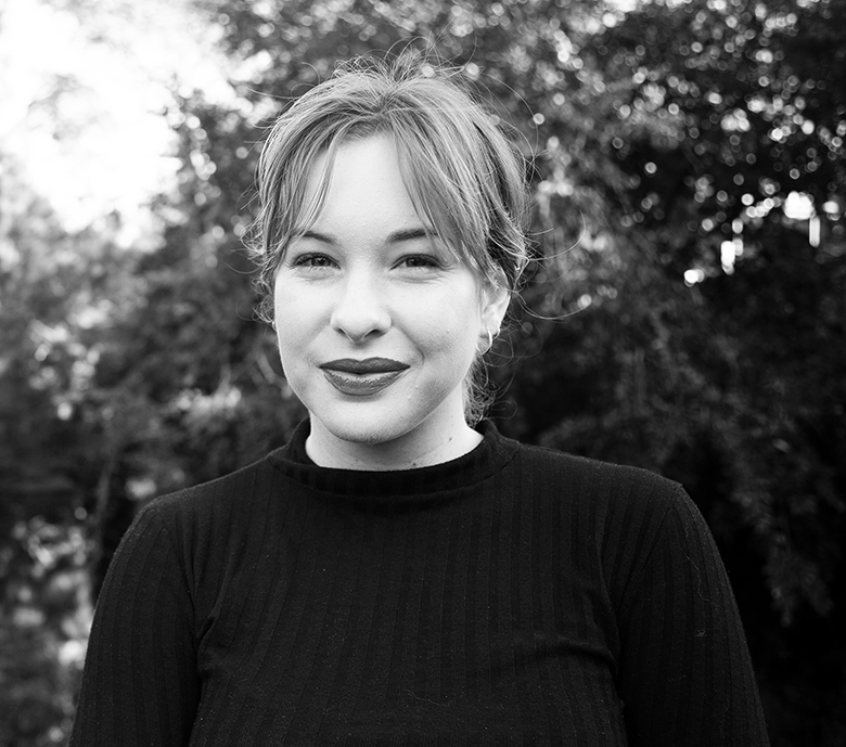 Black and white photo of Alex Philp. She is standing in a garden, wearing a black jumper and she's smiling at the camera.
