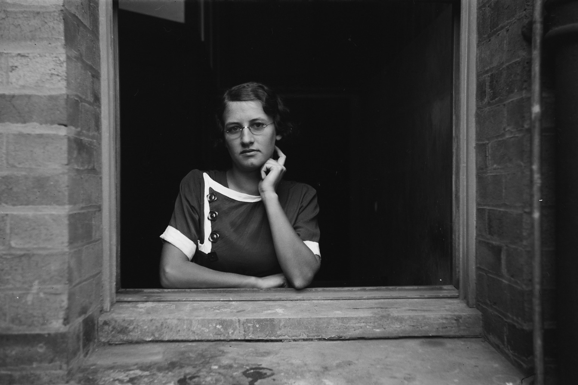 Black and white photograph of a young woman looking out the window of a brick building