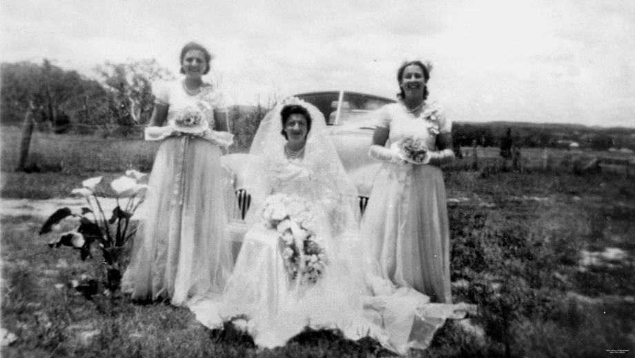 Bride and bridesmaids standing in front of a Holden, ca. 1951. The bride is sat between her two standing bridesmaids wearing a big white bridal gown, holding a bouquet.