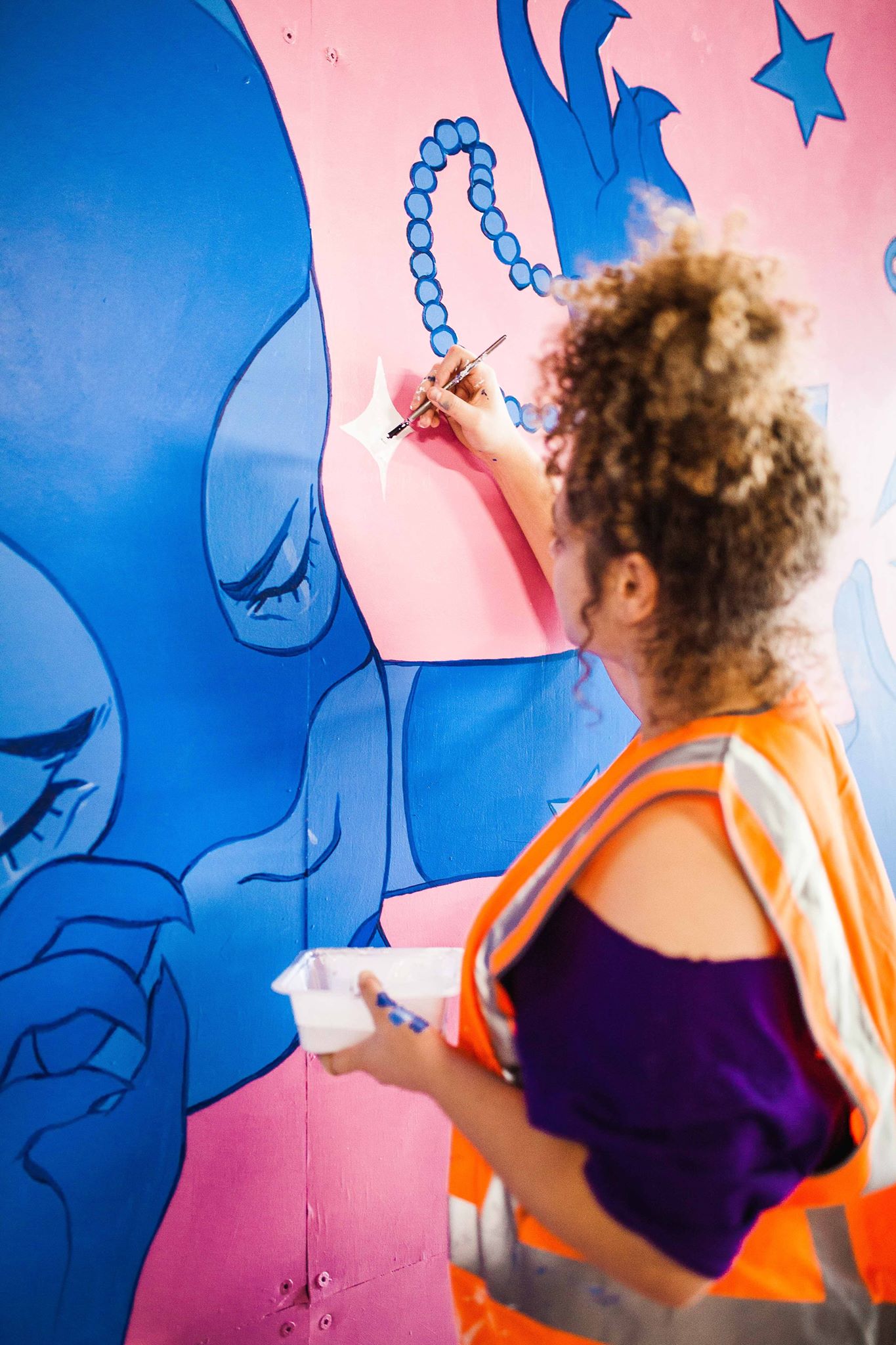 Tori-Jay Mordey wears a high vis vest and a purple shirt. She is standing side on, painting a blue and pink mural