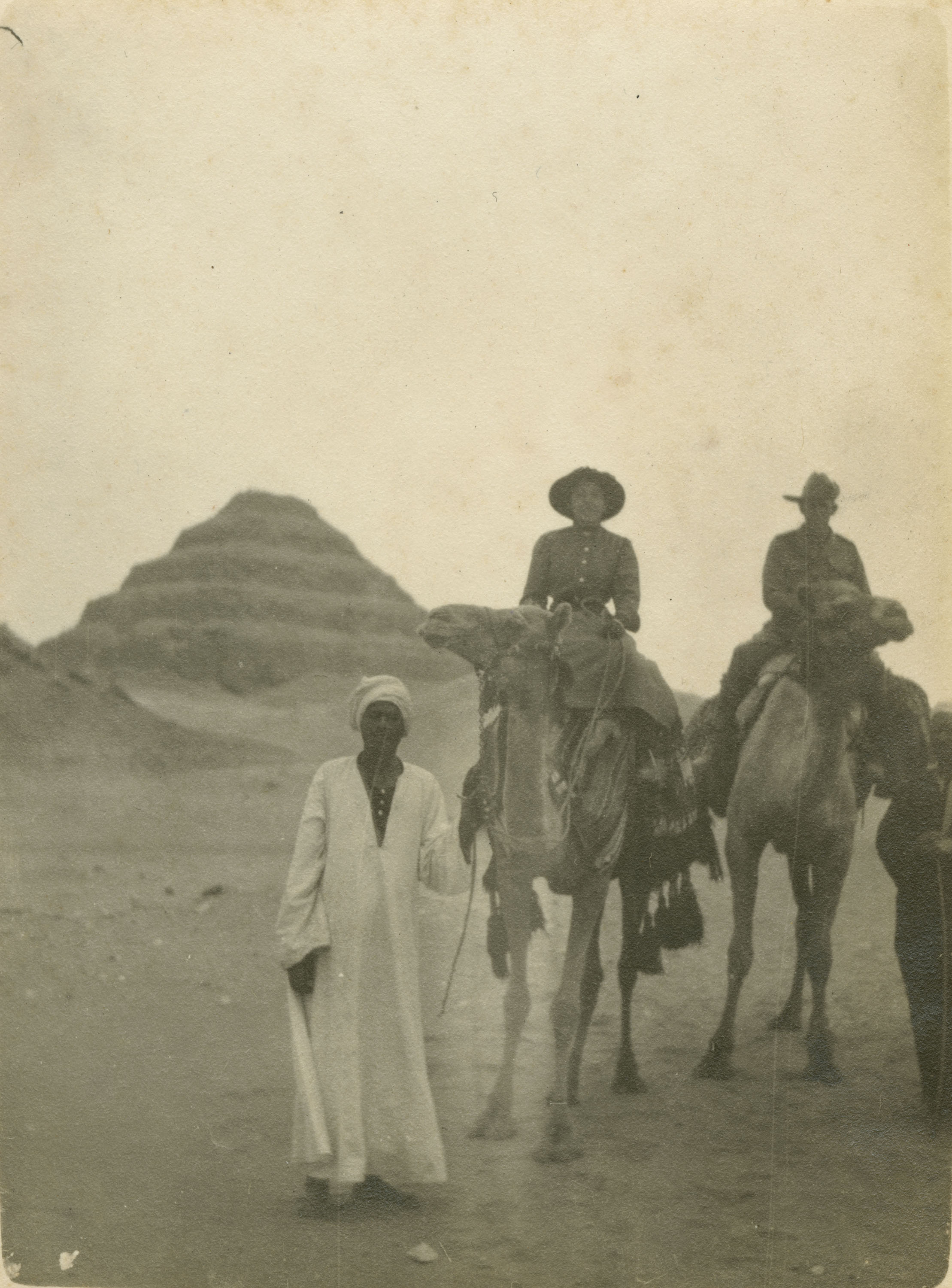 Two people on camels being led by two men with step pyramid in background