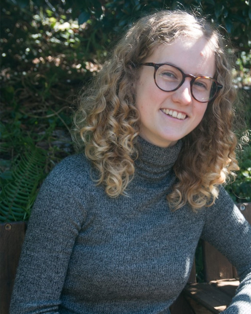 Emily Winter sits at a table in the sunshine. She is smiling, wearing a grey turtleneck and glasses.