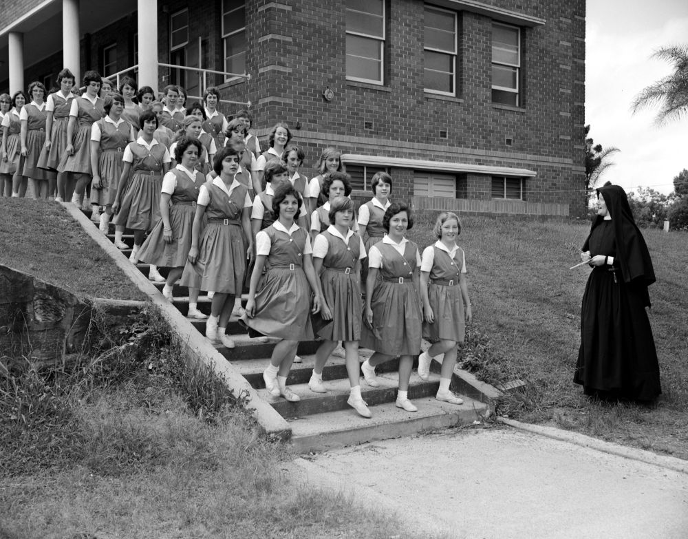 Schoolgirls descending a flight of stairs outside a school building, supervised by a nun in a black habit and veil