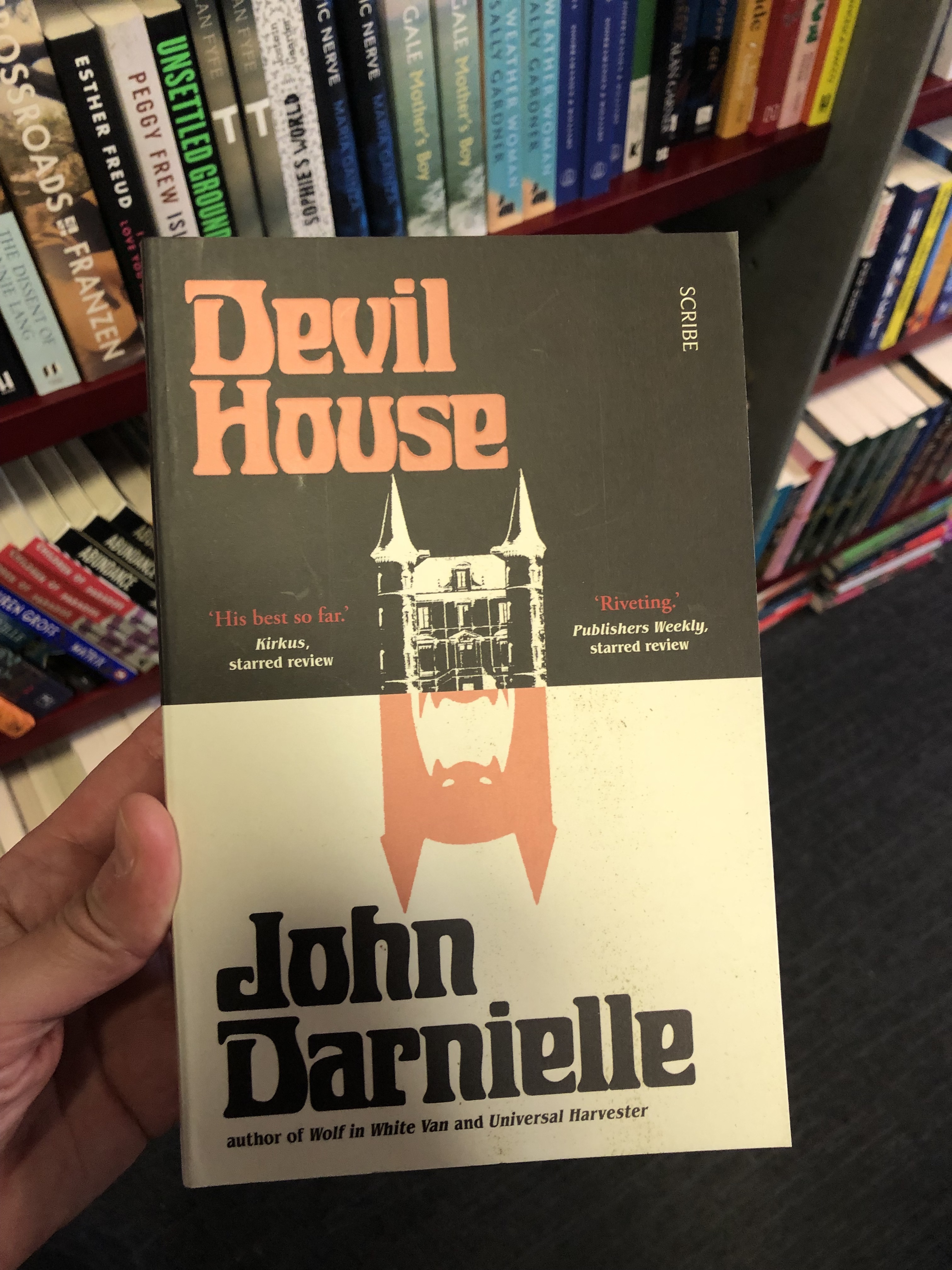 A hand is holding a copy of the book Devil House by John Darnielle. In the background are bookshelves.