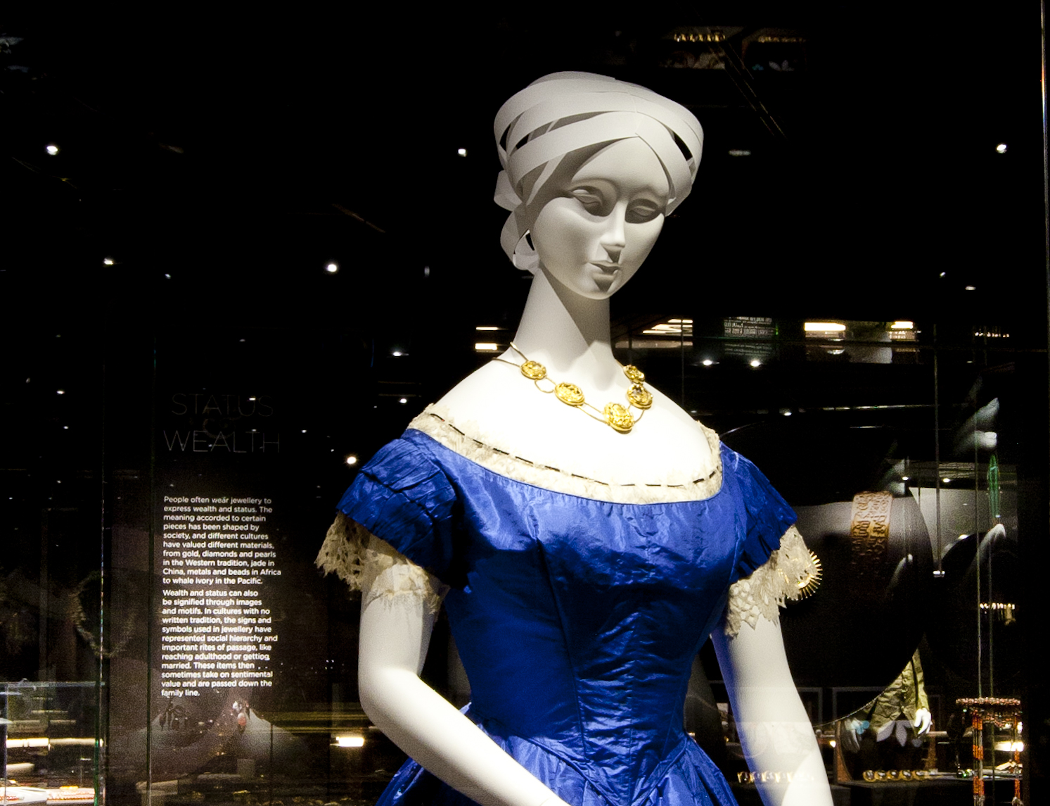 Mannequin in a blue dress wearing the John Watts Necklace as seen in the 2014 exhibition A Fine Possession: Jewellery and Identity. The necklace is an 18 carat gold swag necklace with five openwork carved gold hinged pendants. 