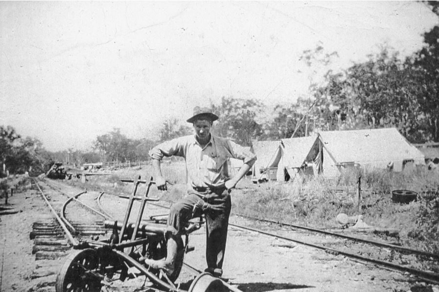 Black and white photo of a railway worker leaning on his trolley on the railway line, with tents in the background