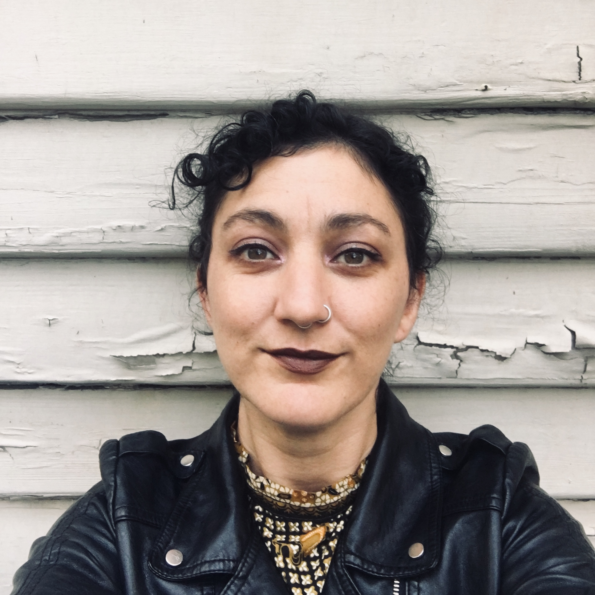 Mykaela Saunders takes a selfie in front of a white wall with peeling paint. She wears a black leather jacket and dark lipstick.