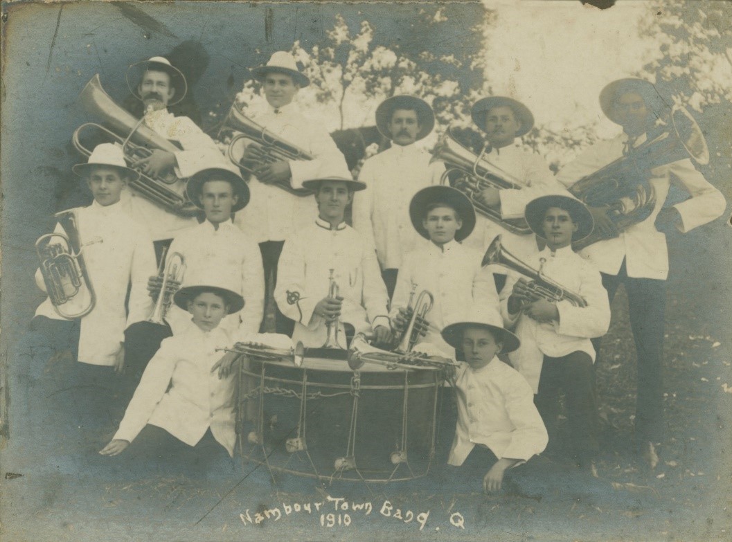 Sepia image of 12 brass band members kneeling and standing around a large drum. Some are holding brass instruments.