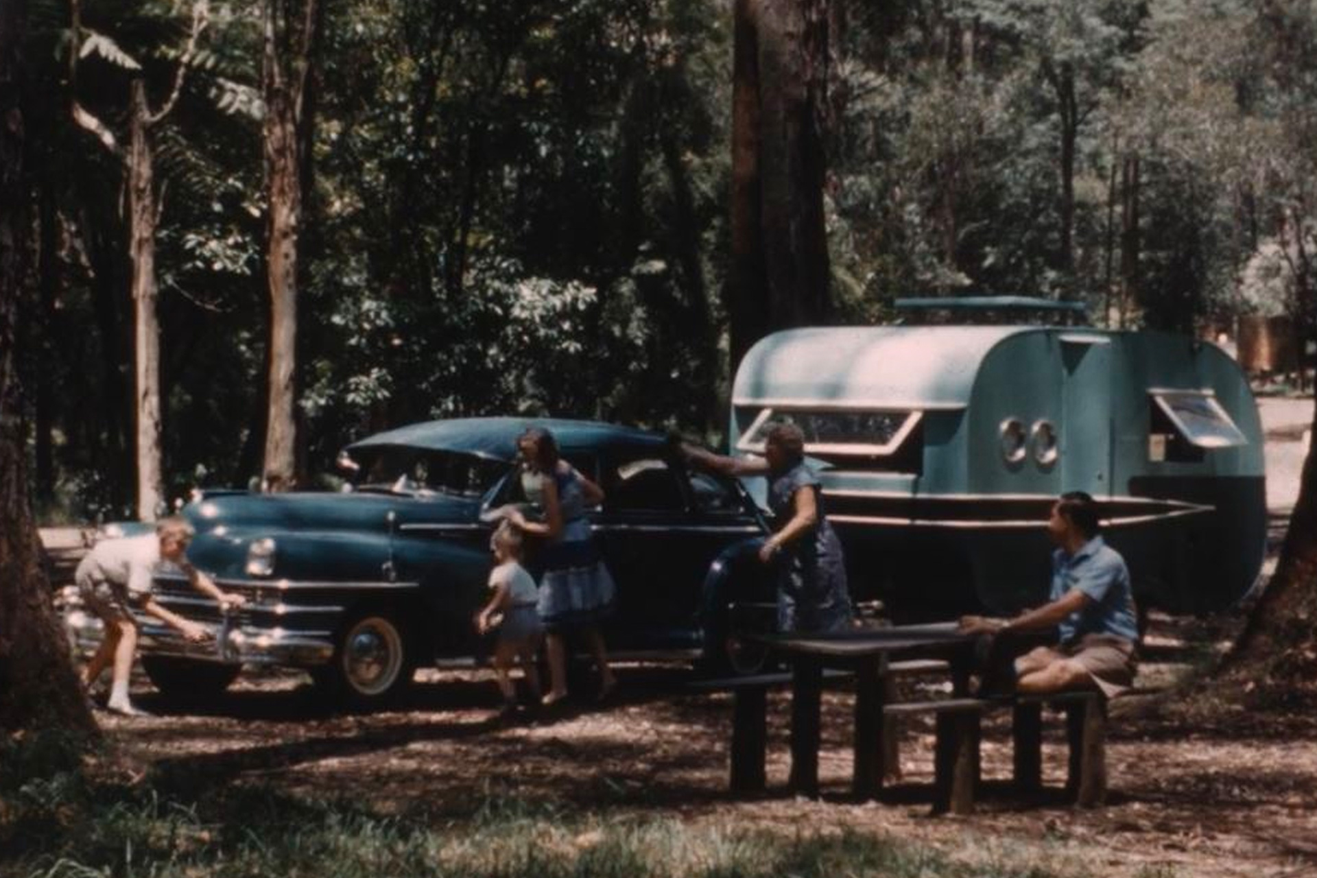 A frame from a home movie, in colour, where a family cleans and polishes a car while the father looks on