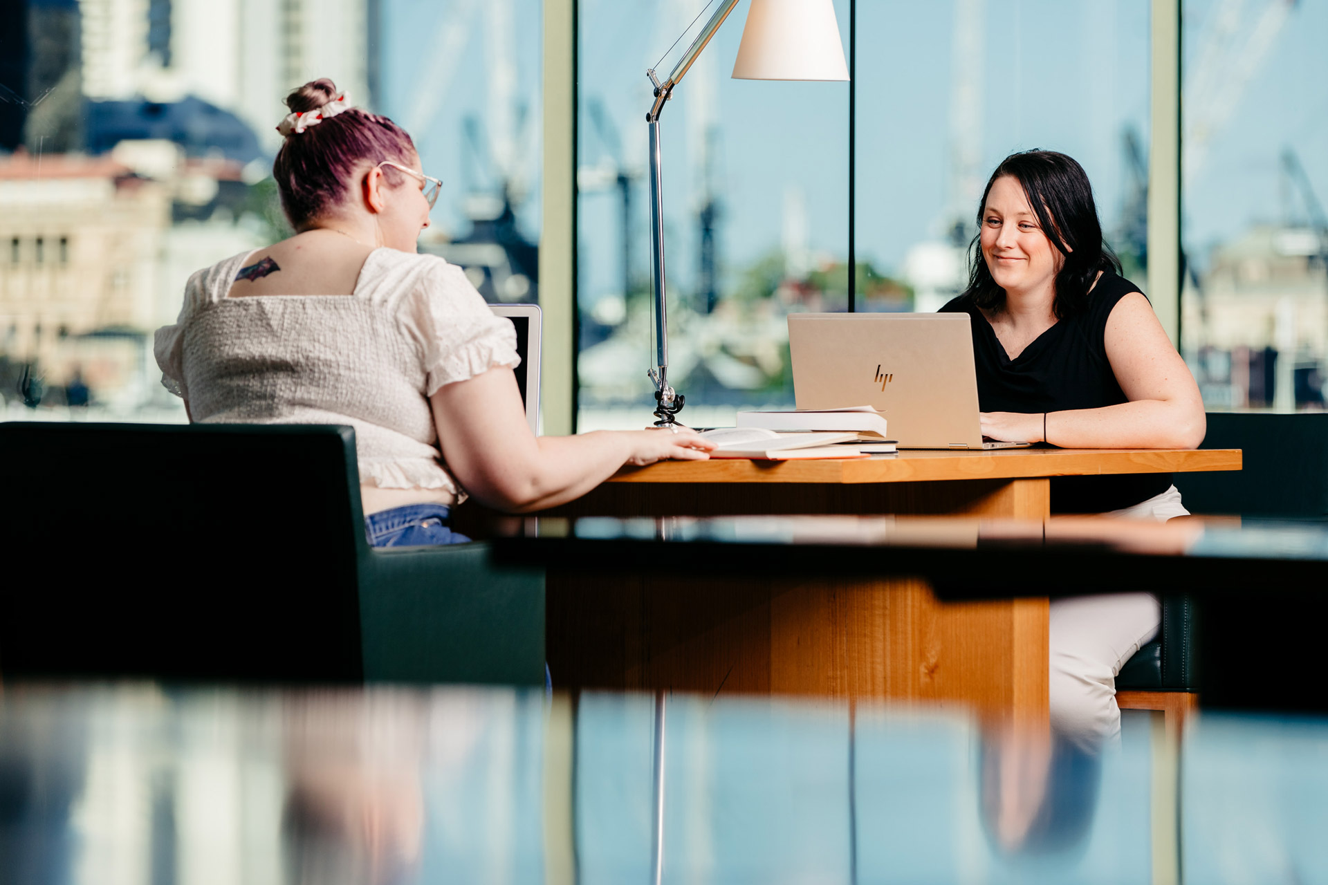 Two women sitting at a joint desk with a view of Brisbane city using laptops at State Library.