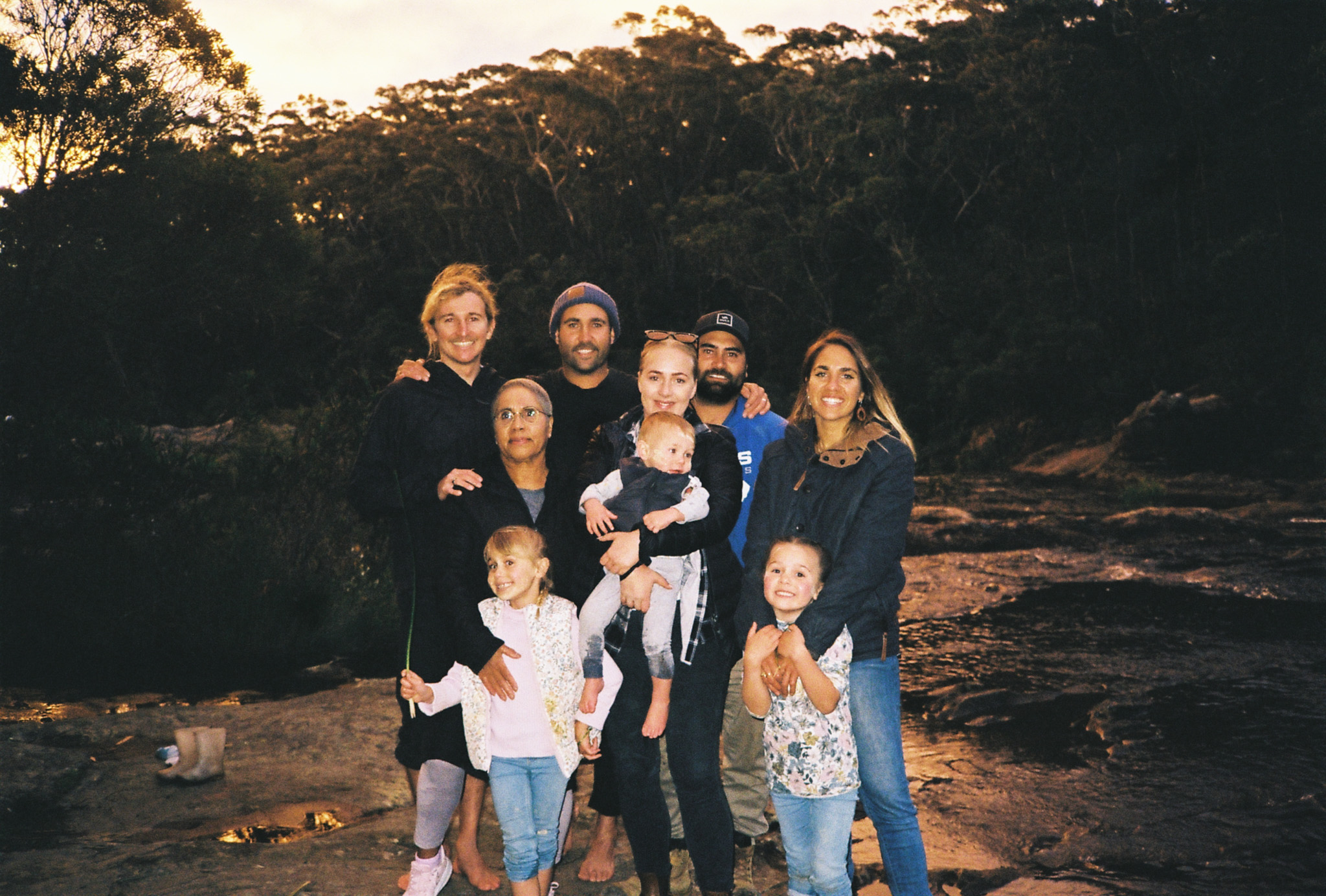 A family stands on rocks with bush in the background. It is sunset. Six adults and three children smile at the camera.