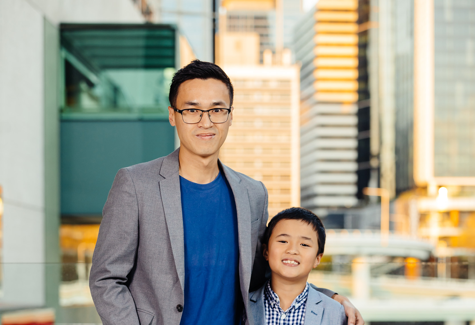 Siang Lu stands in a blue shirt and blazer with his arm around his son. Behind them is skyline of Brisbane at sunset. them 