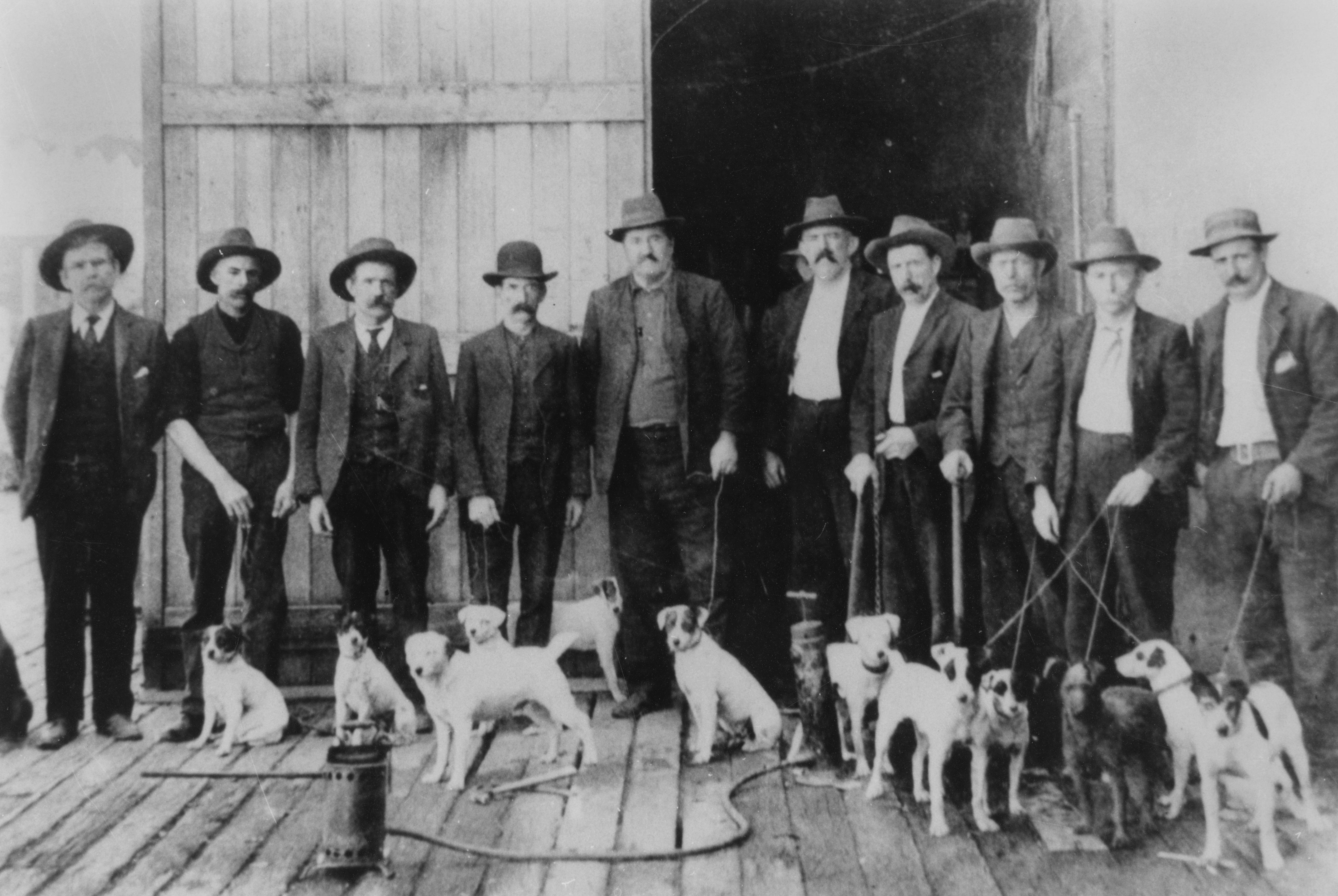 A line of men standing with their dogs outside large wooden doors. 