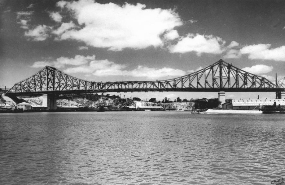 Black and white photo from nineteen fourty showing a distant view of a newly completed Story Bridge. Clouds are in the sky and the sun is shining on the river.