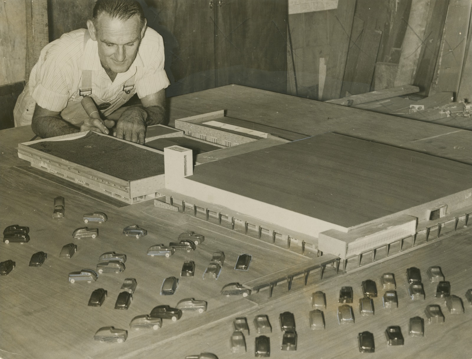 A man leans over a scale model of a department store, complete with miniture cars in the car park.