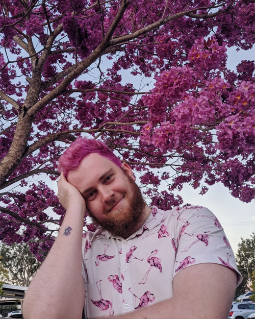 Headshot of Sean West – he wears a white button up shirt with flamingoes on it. His hair is pink and he sits smiling under a pink tree