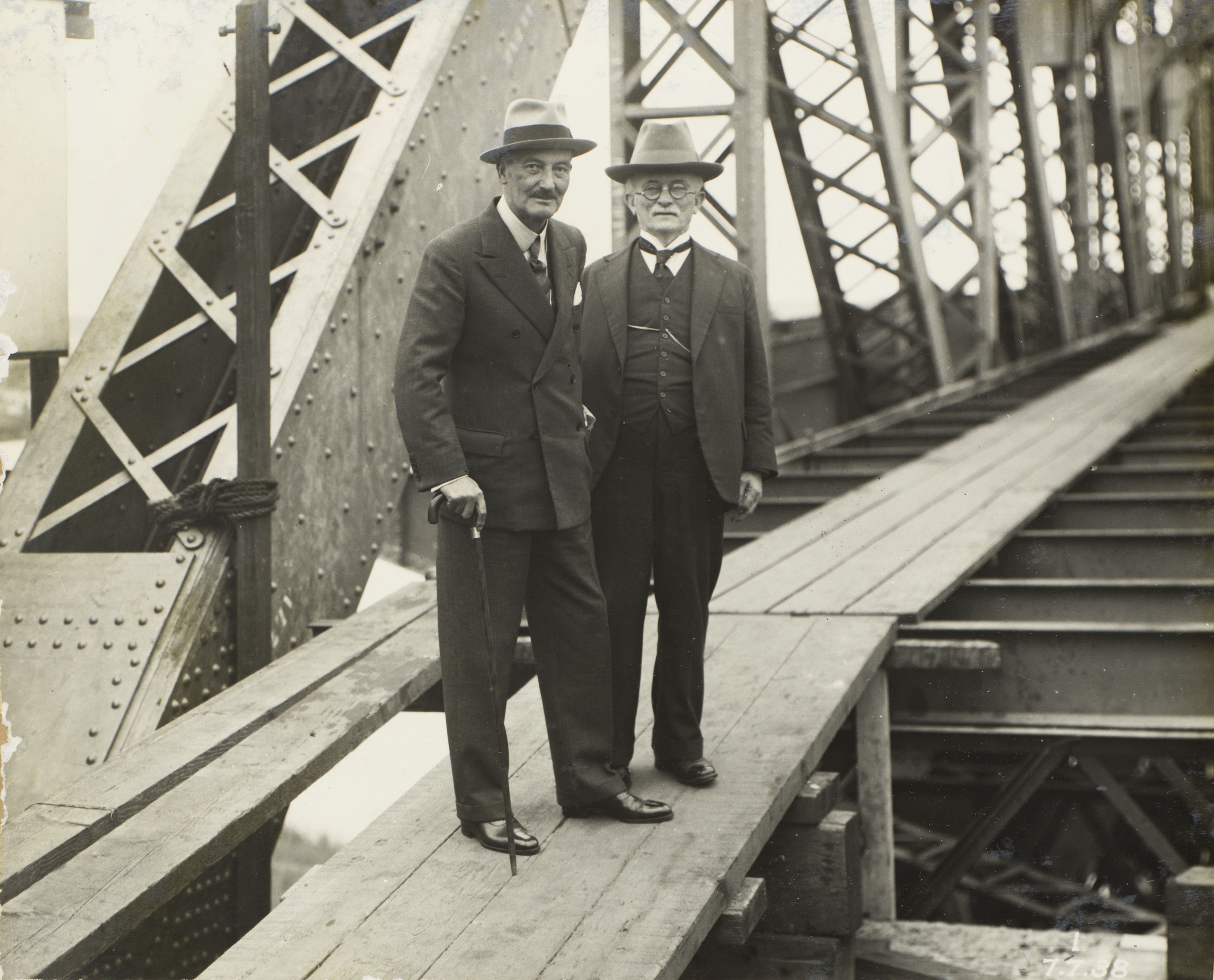 Two men in suits with canes and top hats stand on a makeshift wooden platform spanning the Story Bridge while under construction in 1938