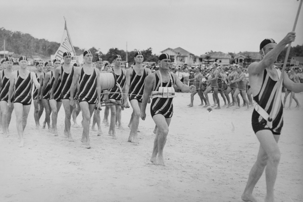 Surf lifesavers marching in striped bathing costumes with flags