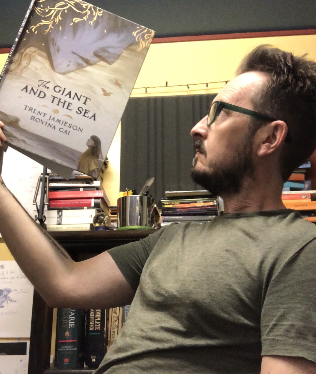 Trent Jamieson holds up a copy of his book, The Giant and the Sea. Behind him are books on shelves