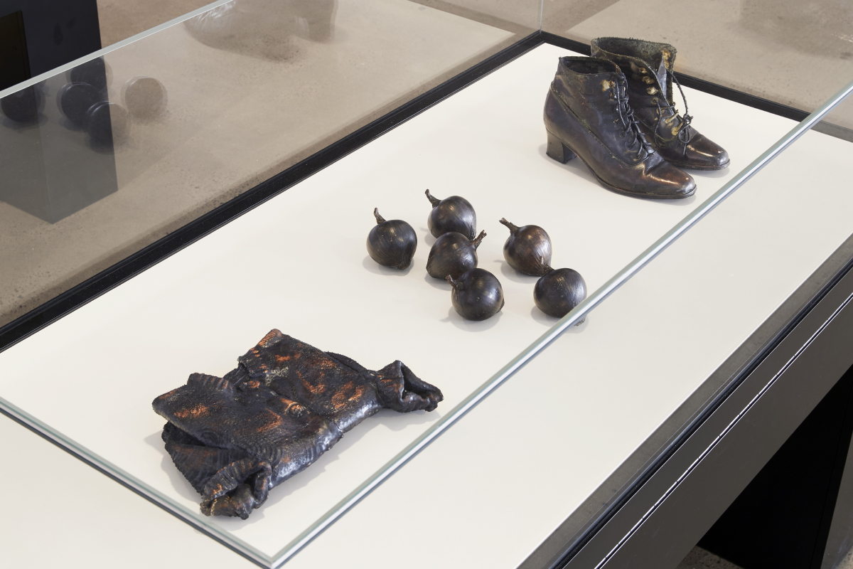 Electro-plated items including folded child's cardigan, 2 garlic bulbs, 4 onions, and a pair of boots in display case.