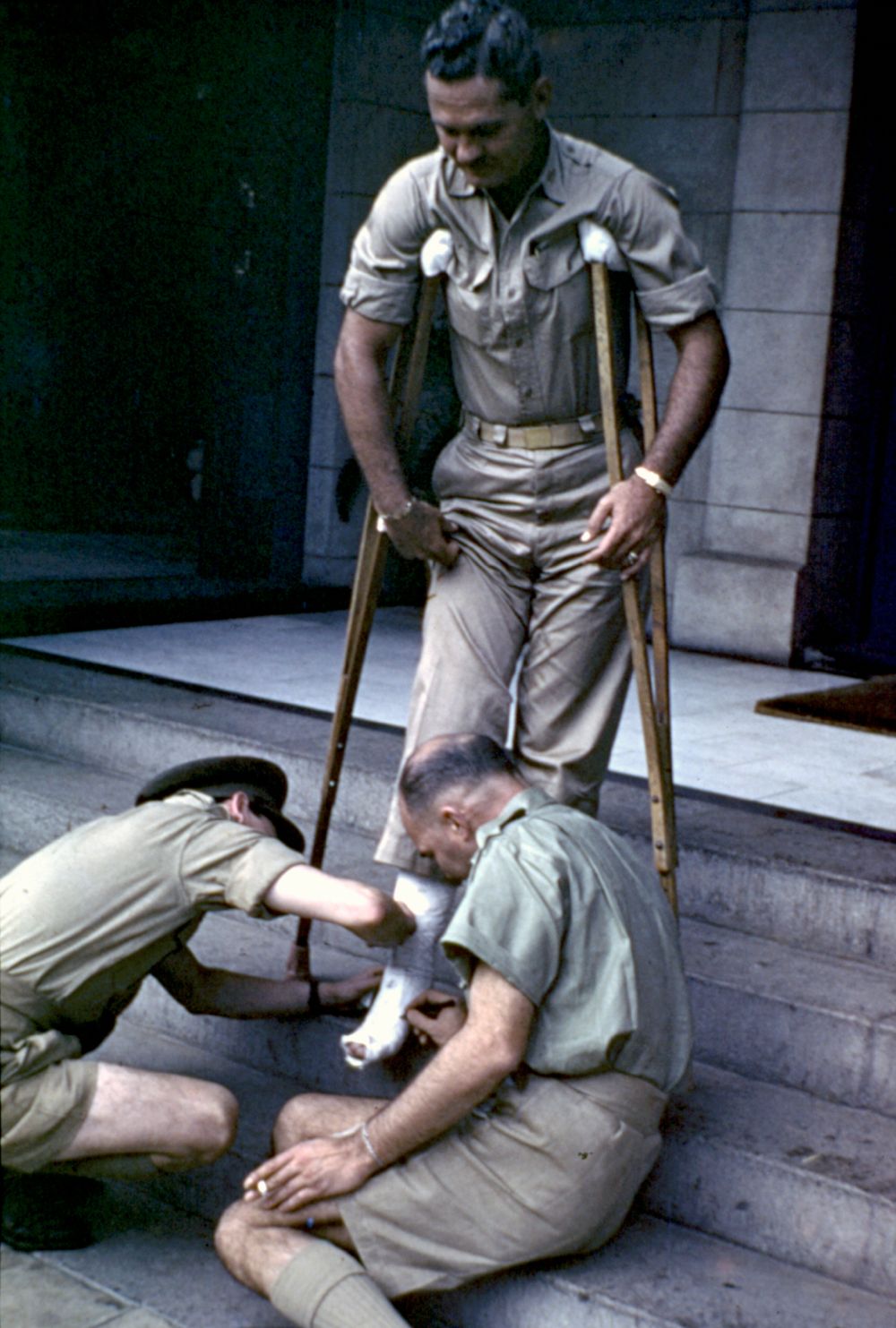 Colour photograph of 2 servicemen sign the leg cast of War correspondent Earl Crotchett on crutches on the steps of the Queens Hotel in Townsville, 1942