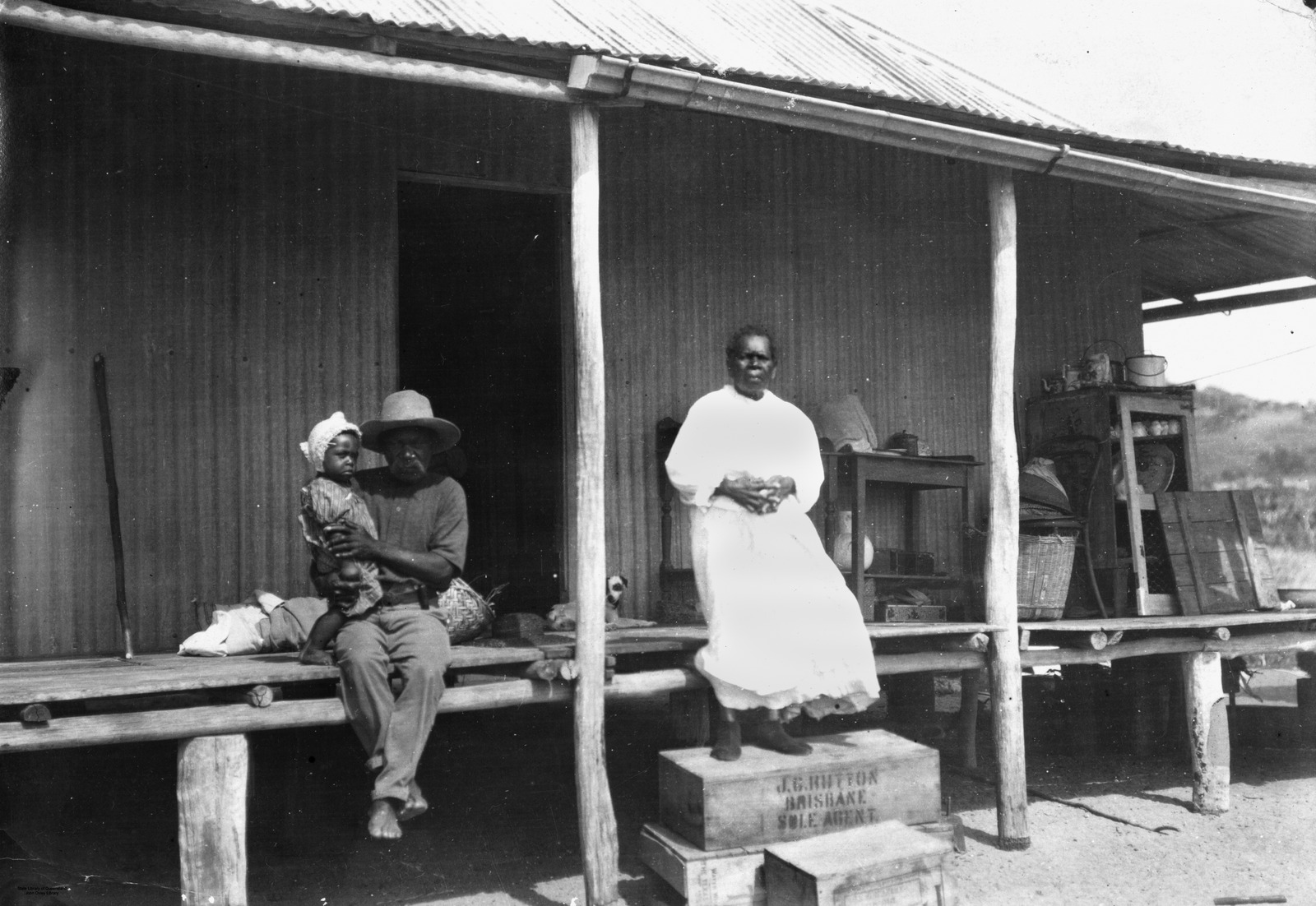 Black and white image of an Aboriginal man sitting holding a child an a woman standing in front of a building in North Queensland, ca.1920