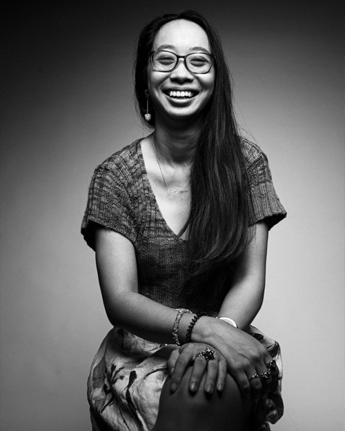 Yen-Rong Wong in a black and white photo. She is seated with her hands folded, smiling, with glasses and her hair long.