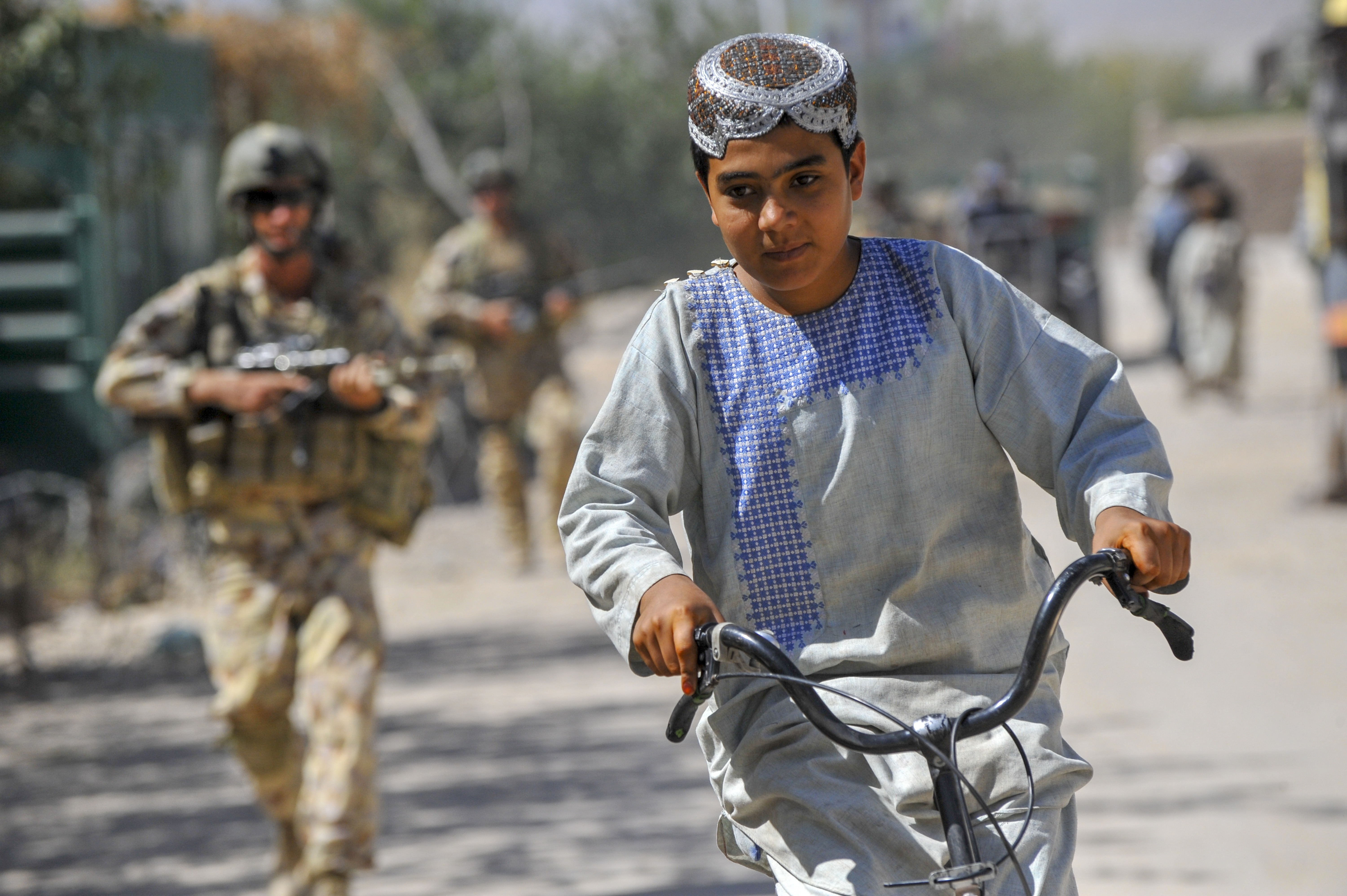 Young Afghan boy rides his bike past Townsville soldiers on foot patrol through the township of Tarin Kowt, Afghanistan, 2009