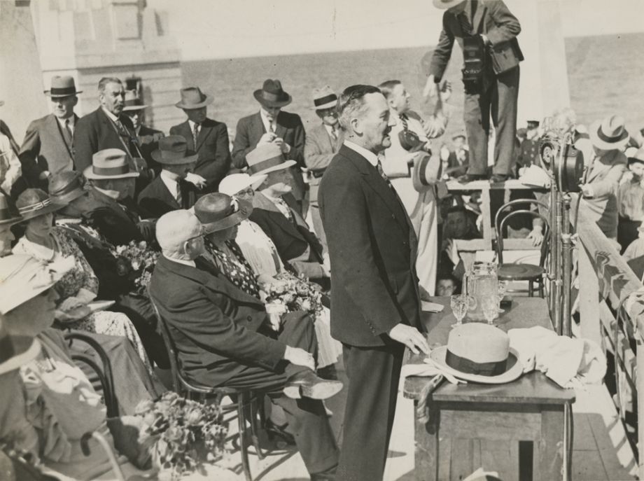 The Governor, Sir Leslie Orme Wilson, stands to make a speech at opening of the Hornibrook Highway, also broadcast on radio. Manuel Hornibrook, his wife Daphne, and other dignitaries sit behind him, with journalists in the background.
