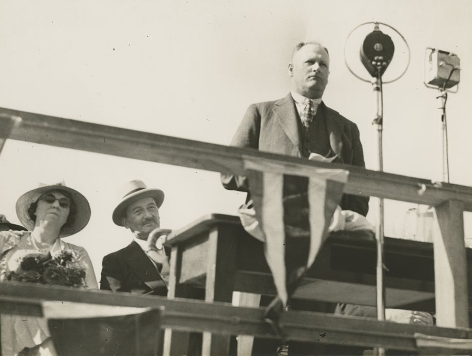 Manuel Hornibrook stands before a large microphone to make a speech on opening day of the Hornibrook Highway. The proceedings were also broadcast on radio. Sir Leslie Orme Wilson, Governor, and his wife, Lady Wilson, are seated behind him.