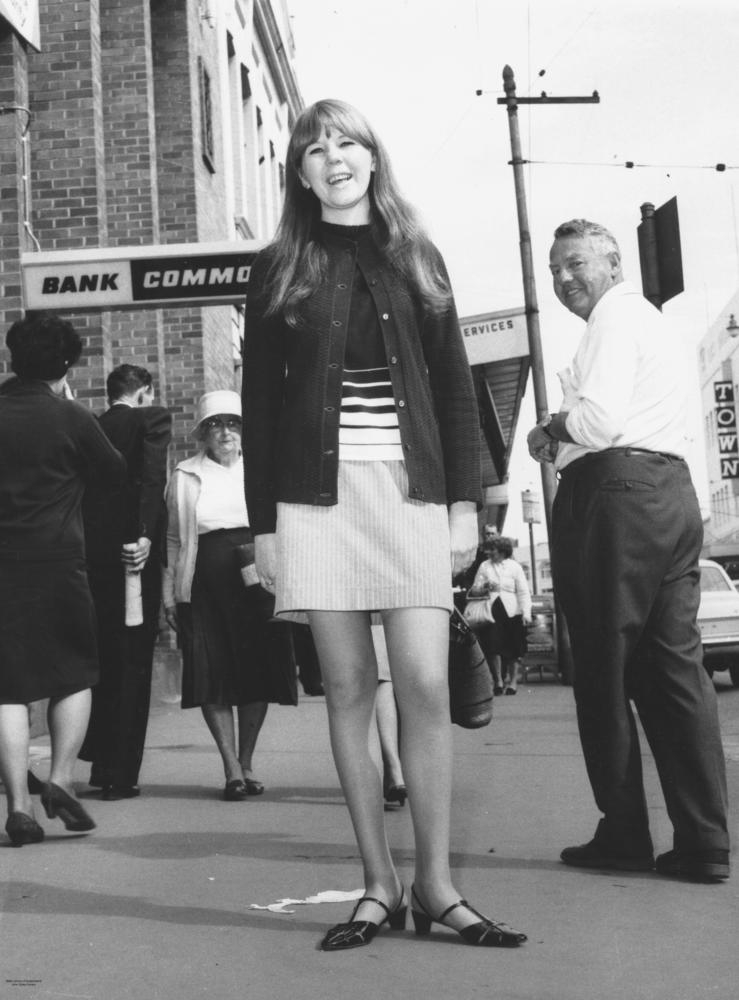Image from 1967 of a young woman smiles into the camera, wearing a casual button up jacket and a mini skirt that reaches her mid thigh. In the background behind her, other people are in formal wear, with women wearing skirts reaching below their knees. A man turns to look at the young woman.