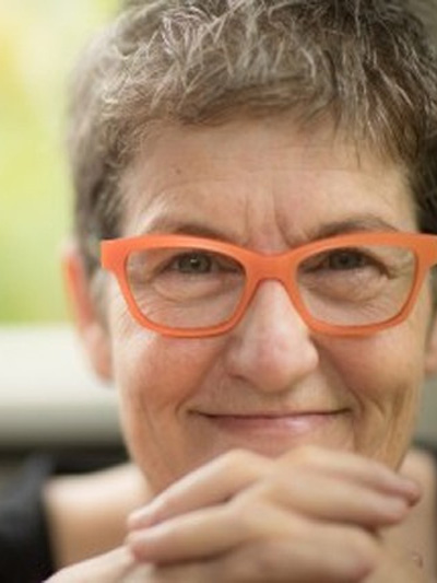 Headshot of Mary-Rose MacColl. The background is blurred green. Mary-Rose wears orange glasses and holds her hands together.