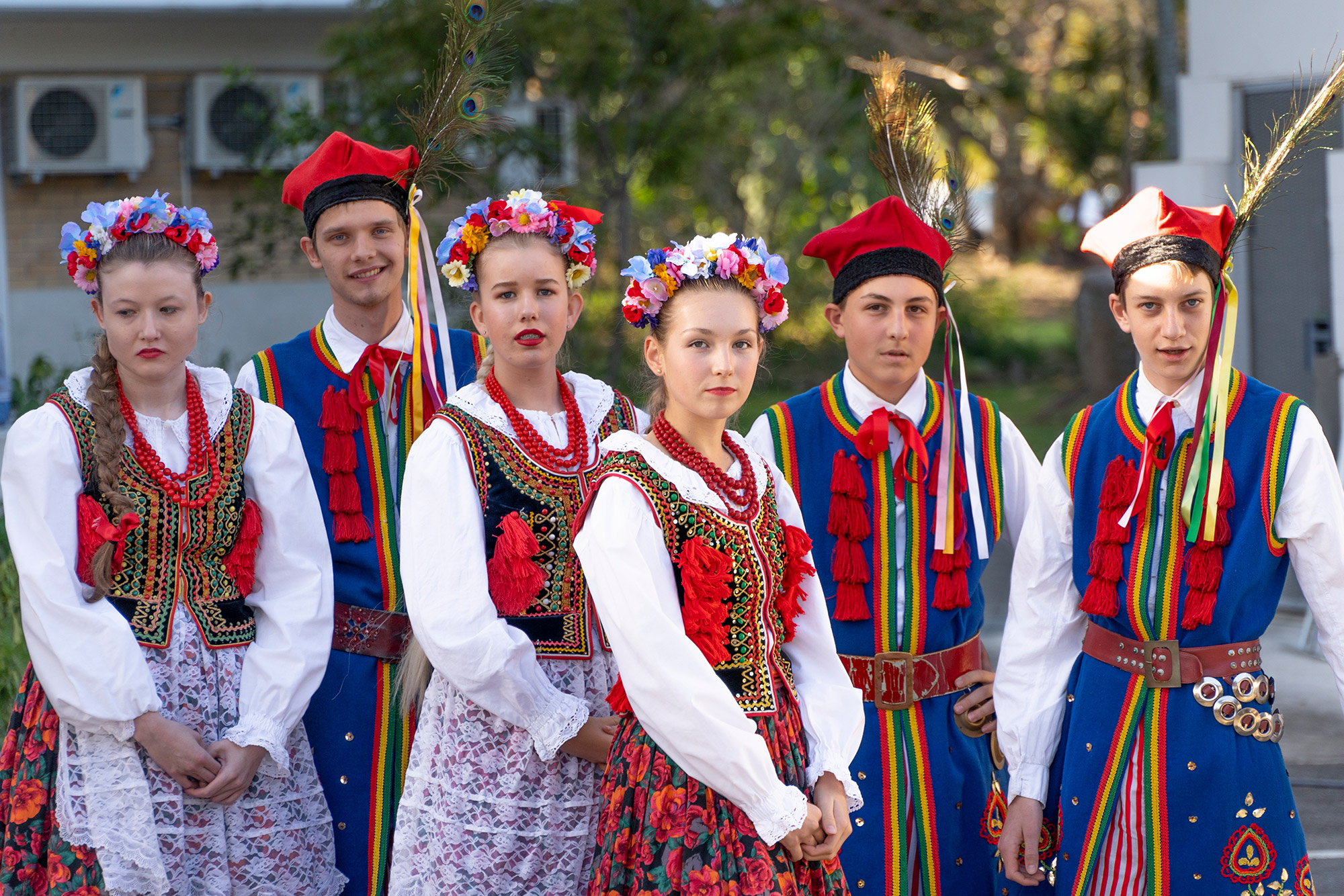 Members of Obertas Polish Folkloric Ensemble waiting to perform. They are dressed in colourful traditional Polish clothes, wearing flower crowns or peacock feathers.