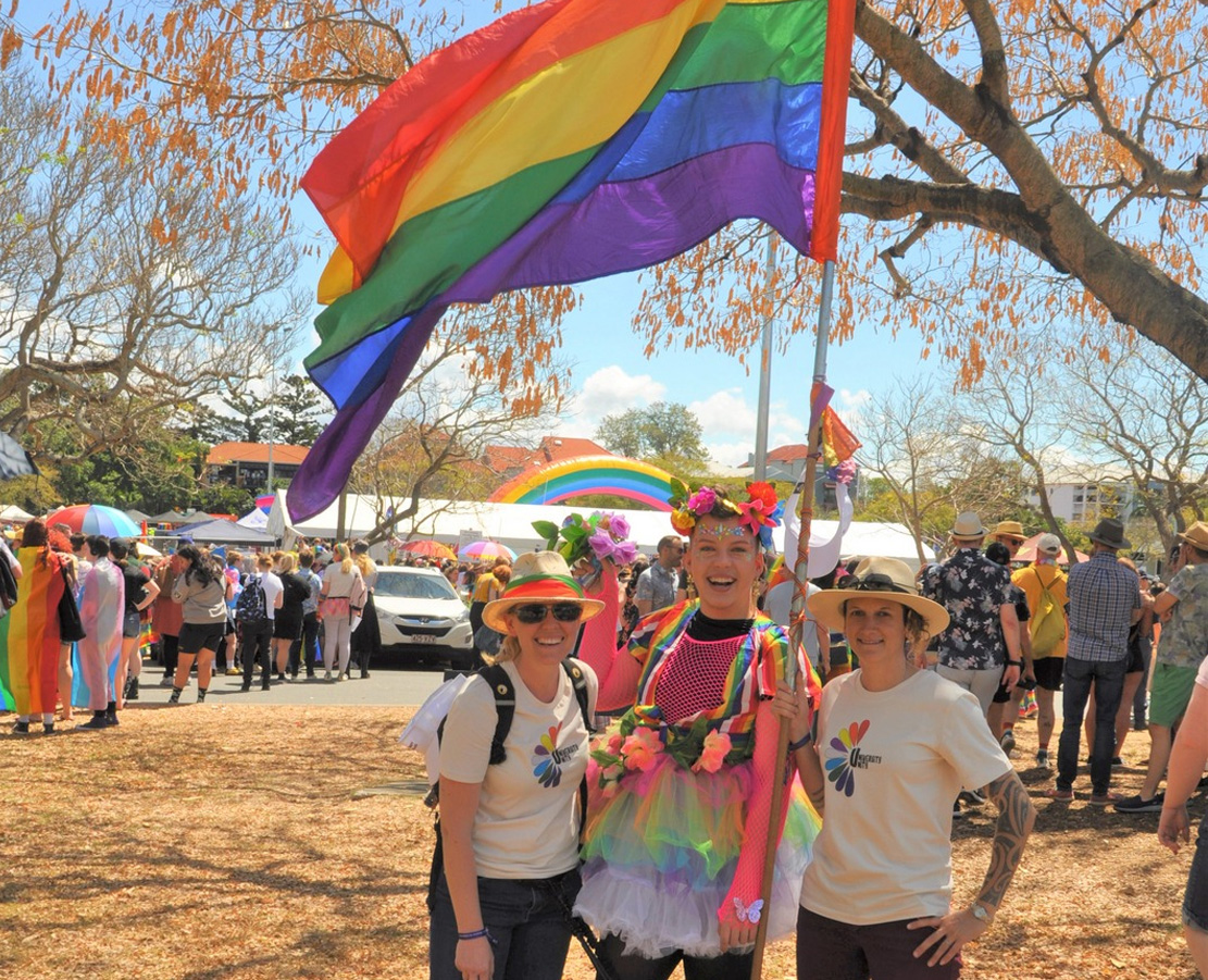 Three women standing together in a park holding a rainbow flag with crowd in background