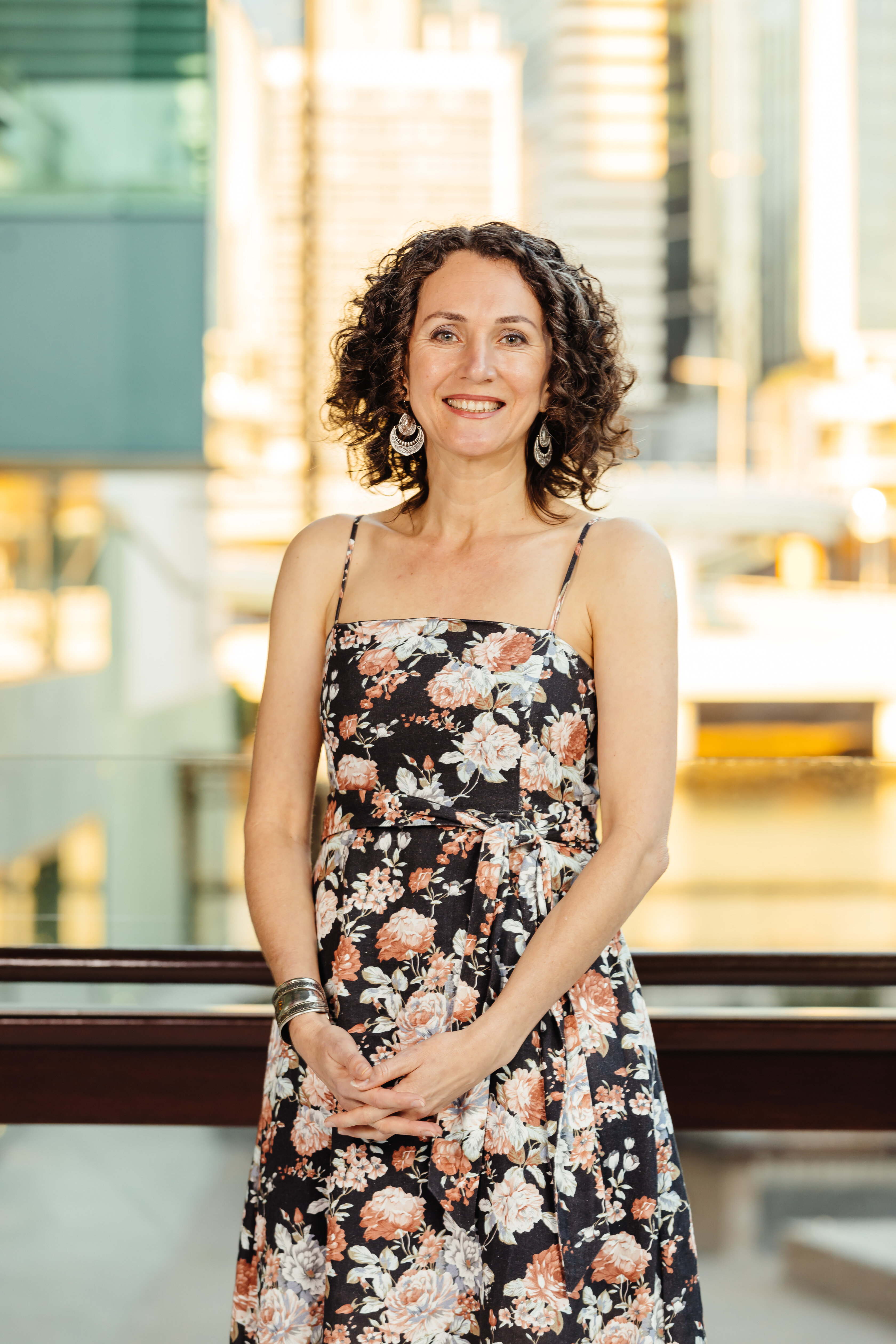 Kali stands at sunset on the Queensland Terrace. She is wearing a black dress with white and brown flowers and is smiling