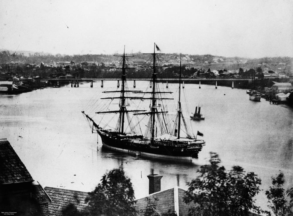 Black and white photo of 3 masted vessel 'Indus' on Brisbane river with rooftops in foreground and bridge in background, ca.1874