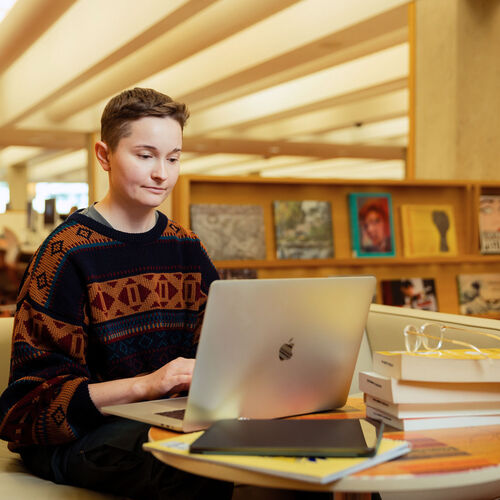 A person sits at a table in the State Library, working on a laptop with a pile of books, with shelves lining the background.